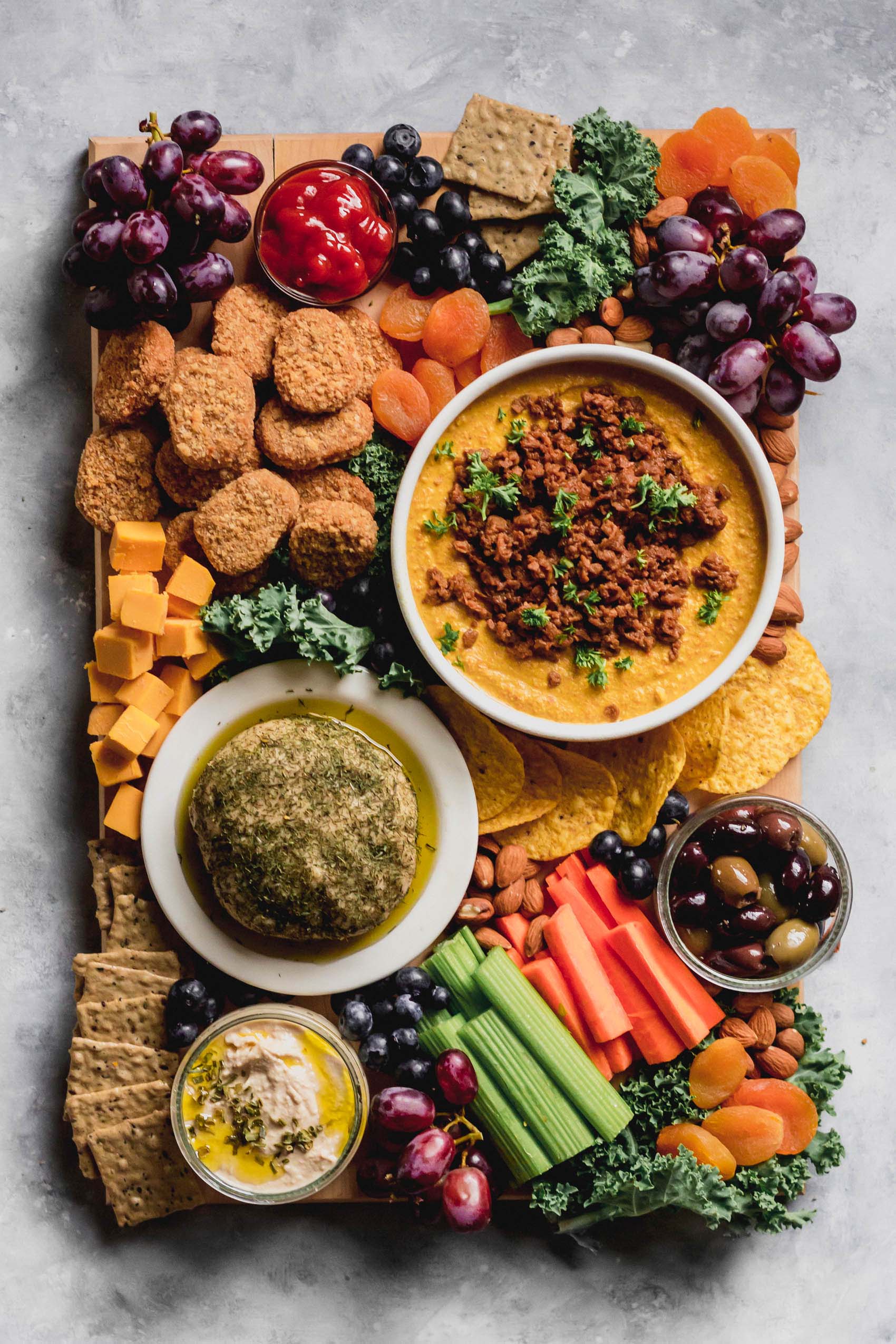 a platter topped with fruit, vegan nuggets, chili topped vegan queso, a vegan cheeseball, crackers and olives