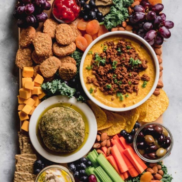 a tray of vegan party foods such as vegan nuggets, cashew queso topped with chili, crackers, fruit and olives