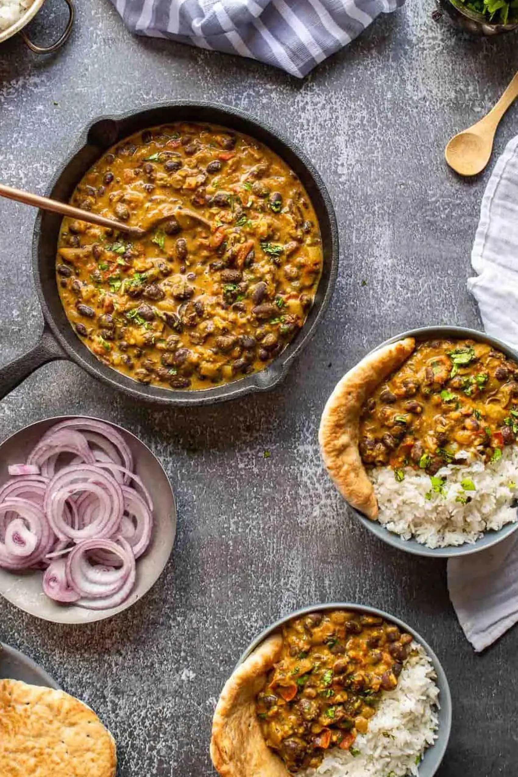 a skillet of black bean curry next to two individual servings and a bowl of marinated red onions