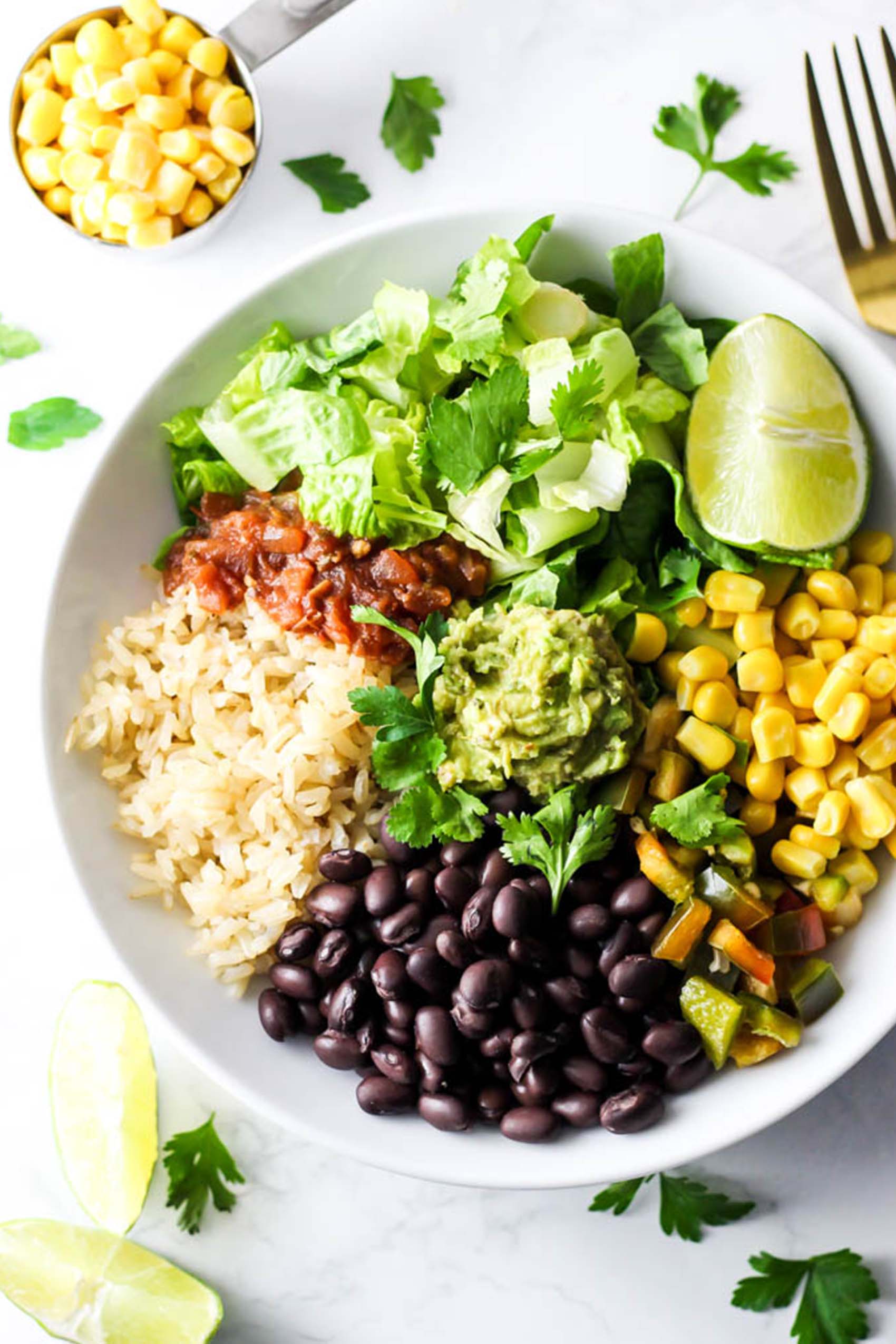 a vegan burrito bowl containing black beans, corn, lettuce, salsa, brown rice, guacamole and a wedge of lime