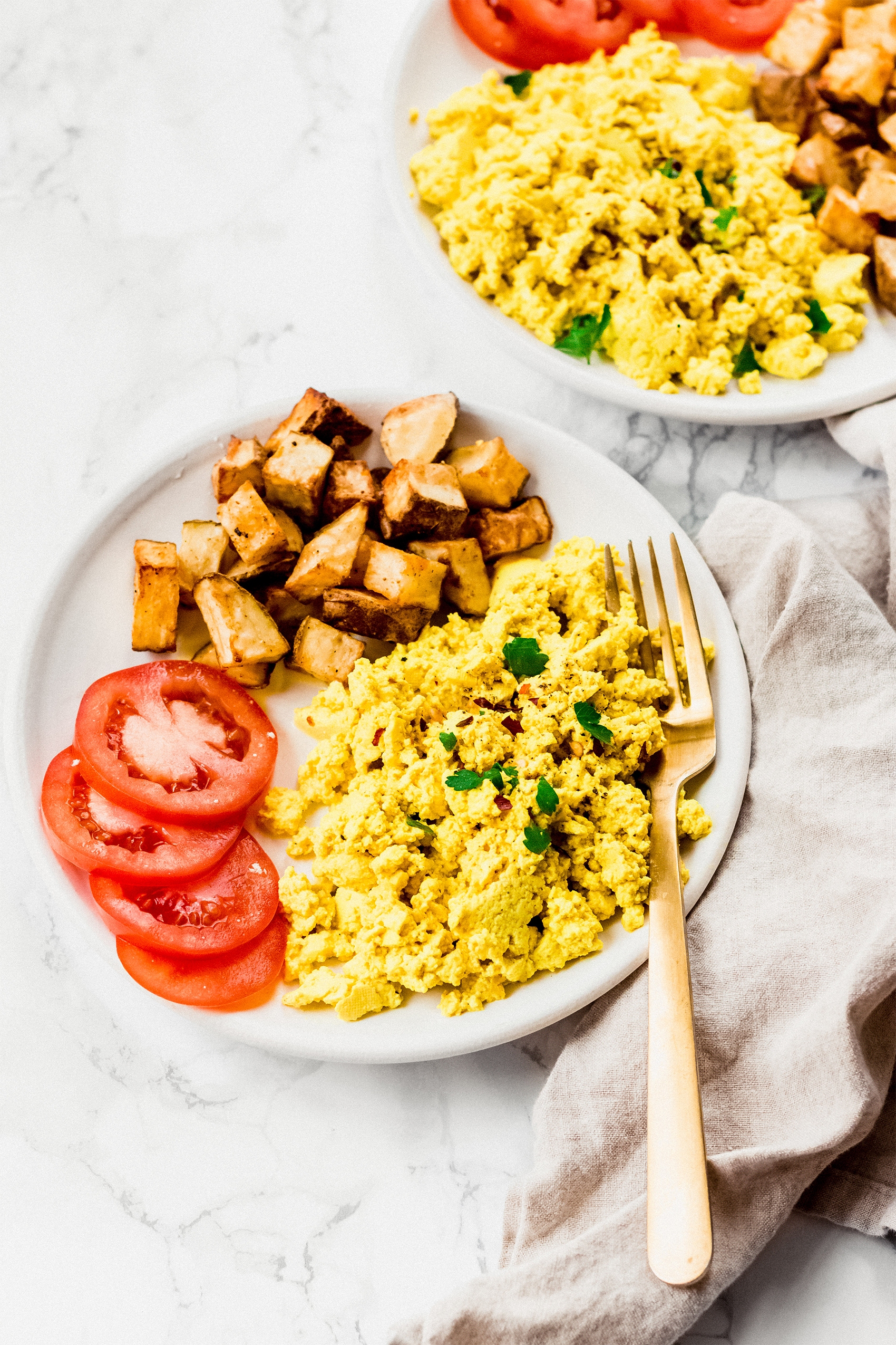 two plates of tofu scramble served with breakfast potatoes and sliced tomatoes