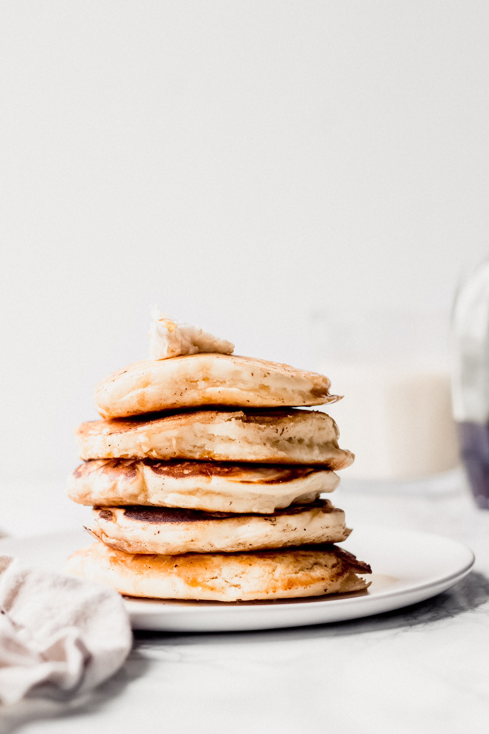 a plate of five pancakes stacked on top of one another, topped with butter and served with a glass of milk in the background