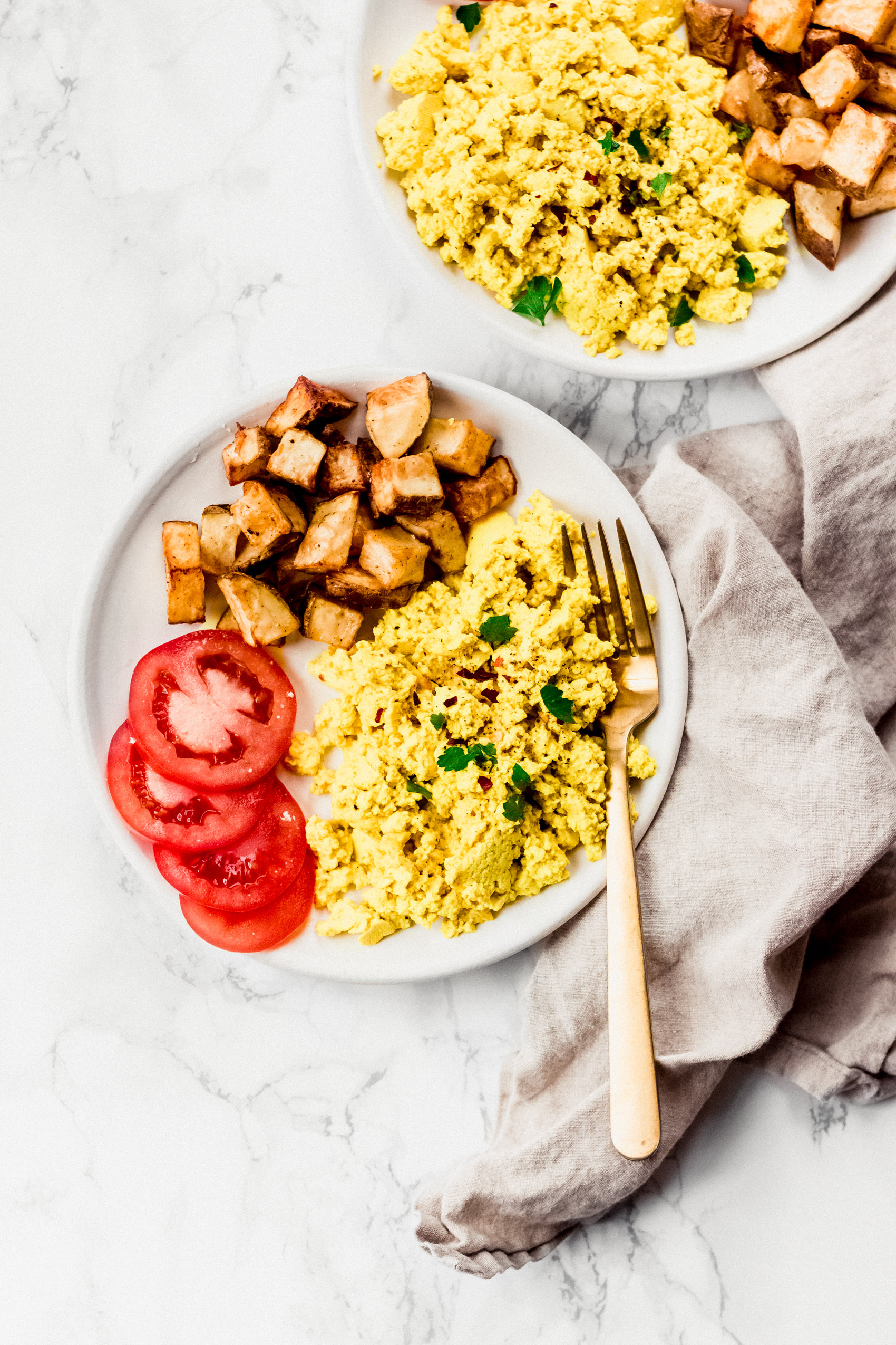 two plates of scrambled tofu with fresh tomatoes and breakfast potatoes