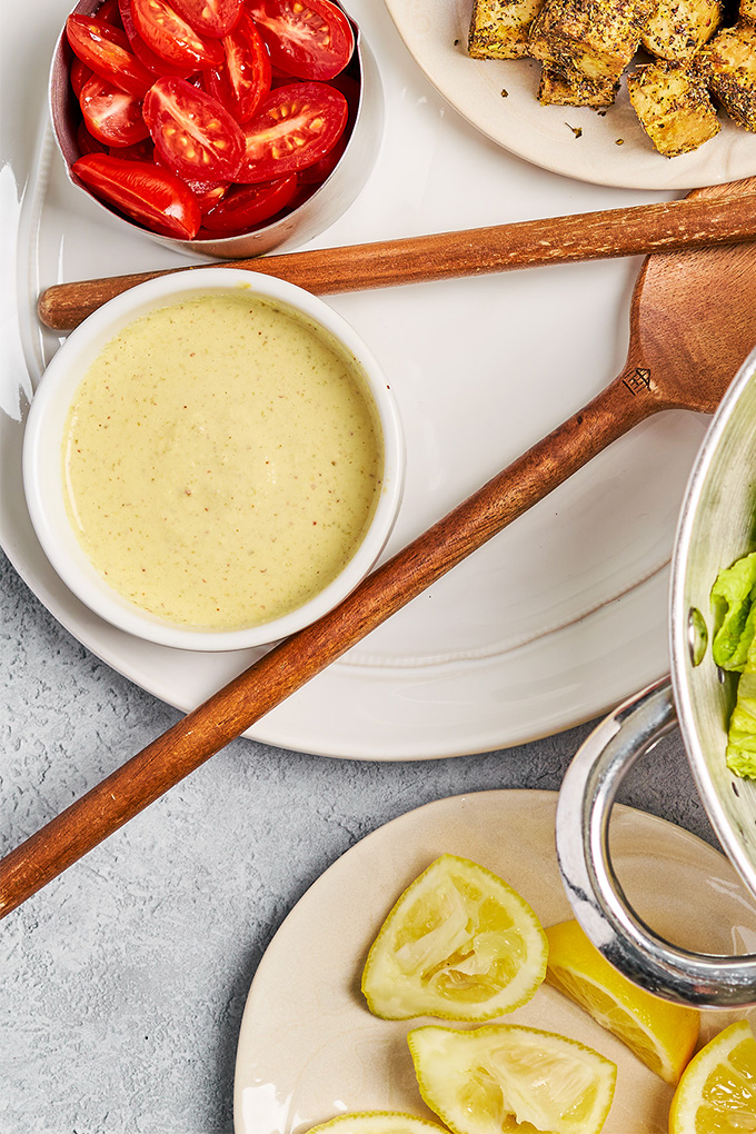 a bowl of homemade vegan caesar dressing next to a plate of lemon wedges and a bowl of lettuce