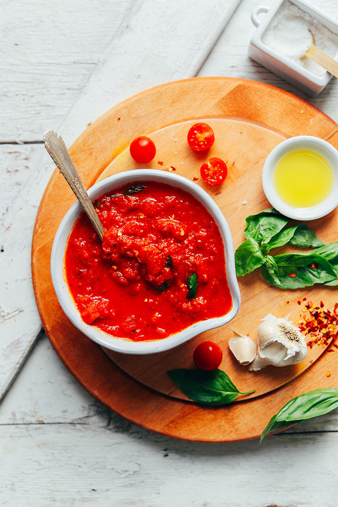 a bowl of homemade marinara sauce surrounded by garlic, basil, olive oil, tomatoes and chili flakes