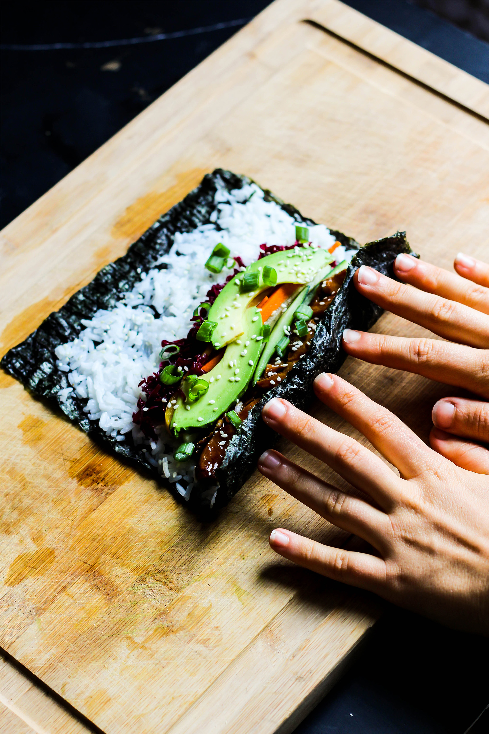 a sheet of nori topped with rice, avocado, and vegetables being rolled into sushi on a cutting board