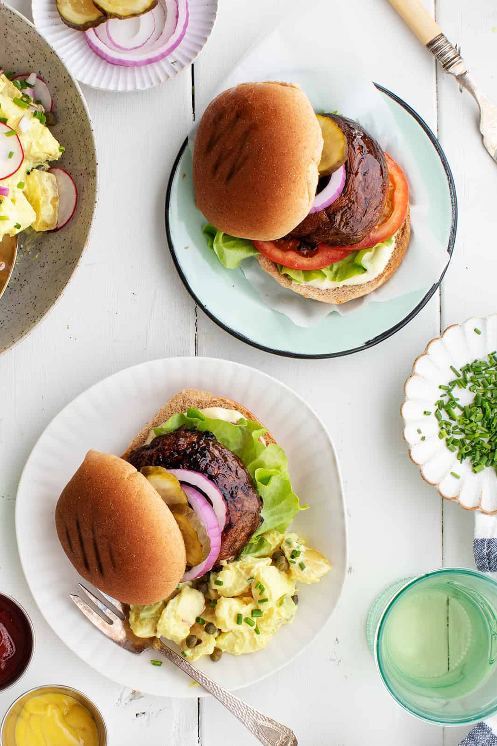 two plates, each with a portobello mushroom burger topped with veggies and served with potato salad