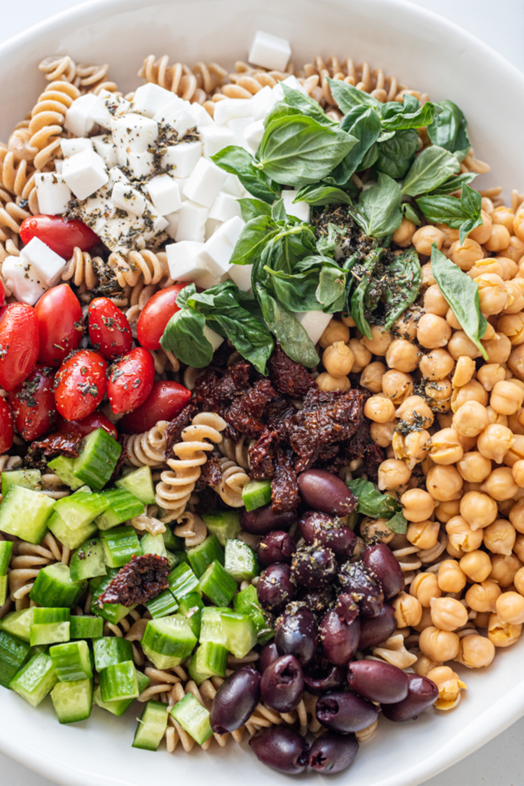 a large bowl containing chickpeas, pasta, olives, cherry tomatoes, vegan cheese and fresh herbs