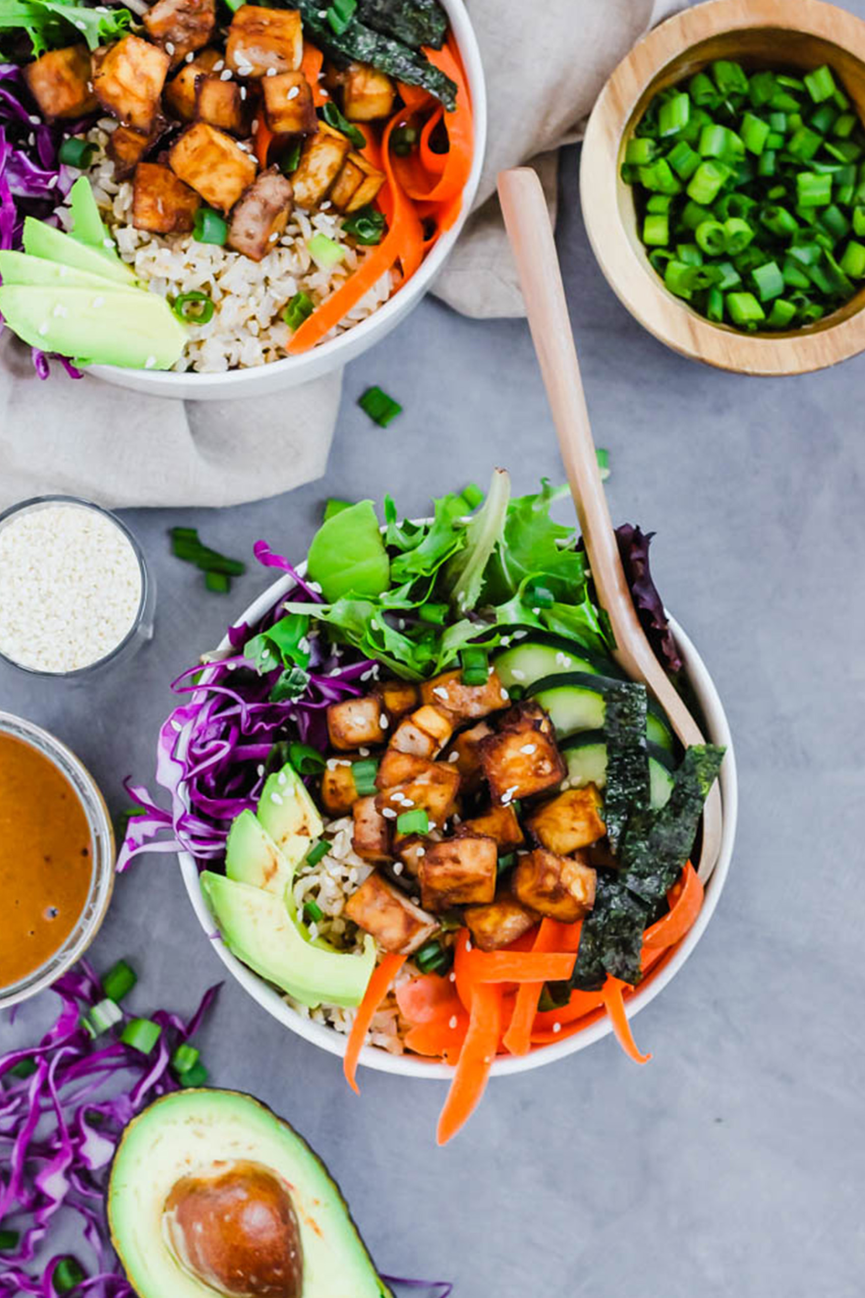 a bowl filled with greens, cucumber, cabbage, carrots, avocado and teriyaki tofu