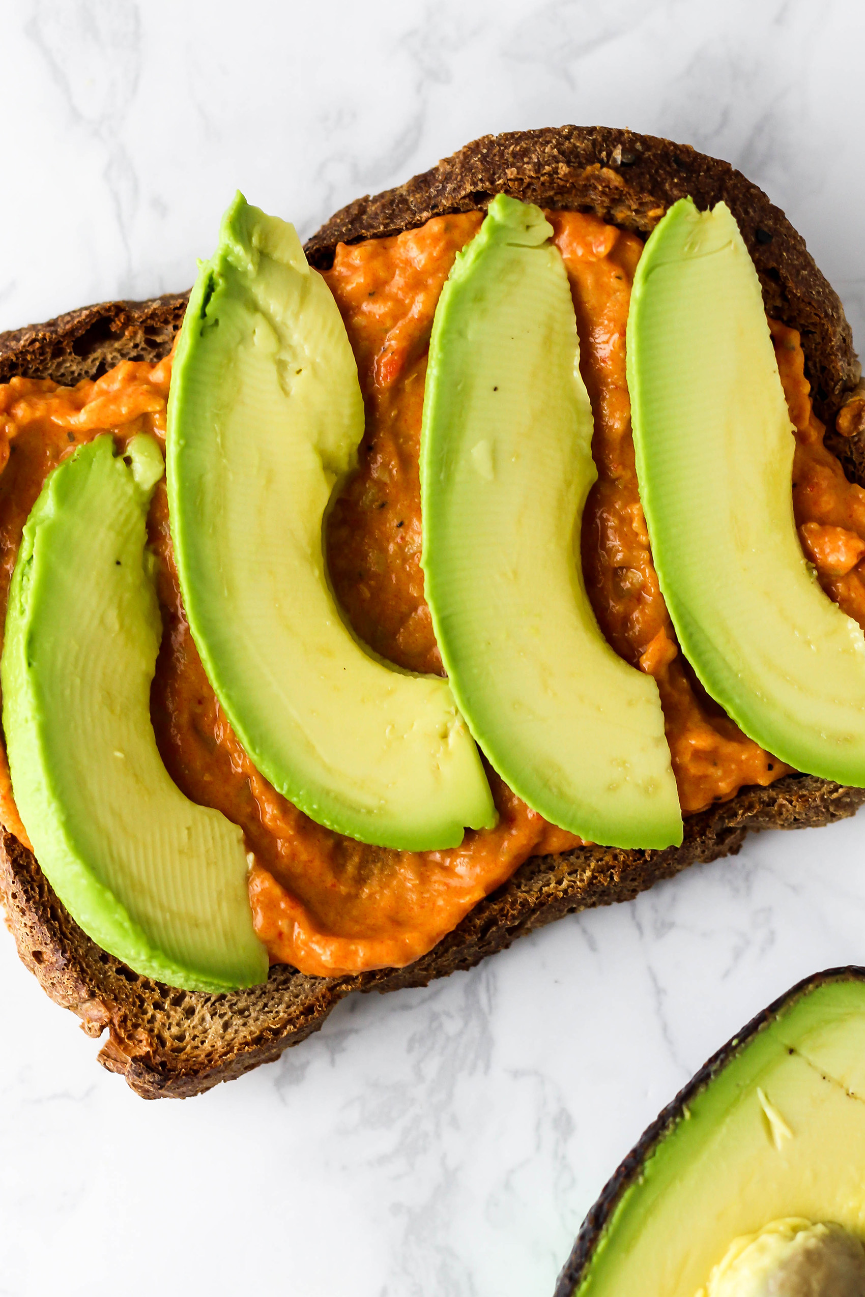 a close up of a slice of toast topped with red pepper hummus and avocado slices