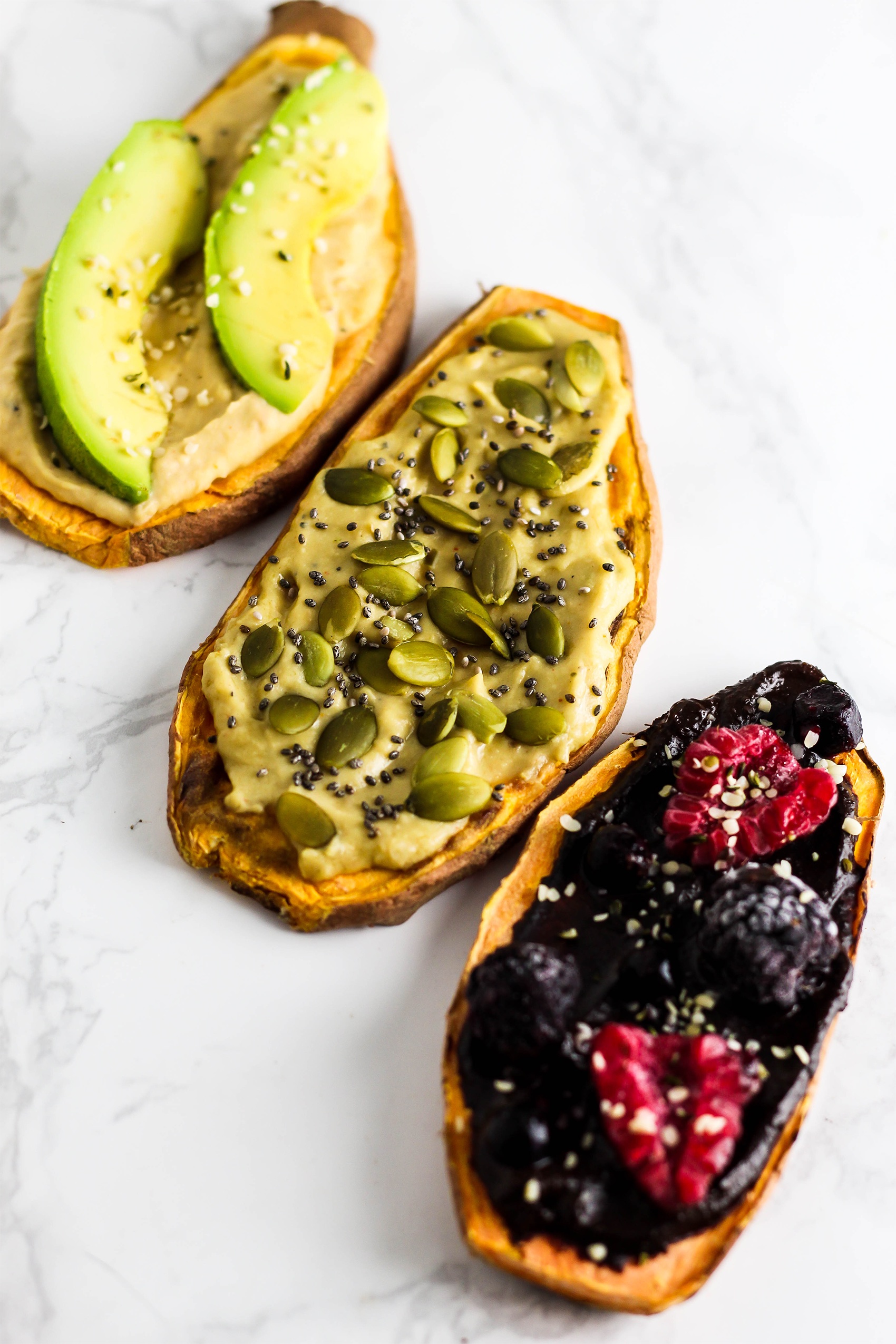 three slices of toasted sweet potato, one topped with hummus and avocado, one topped with hummus and pepitas and one topped with berries