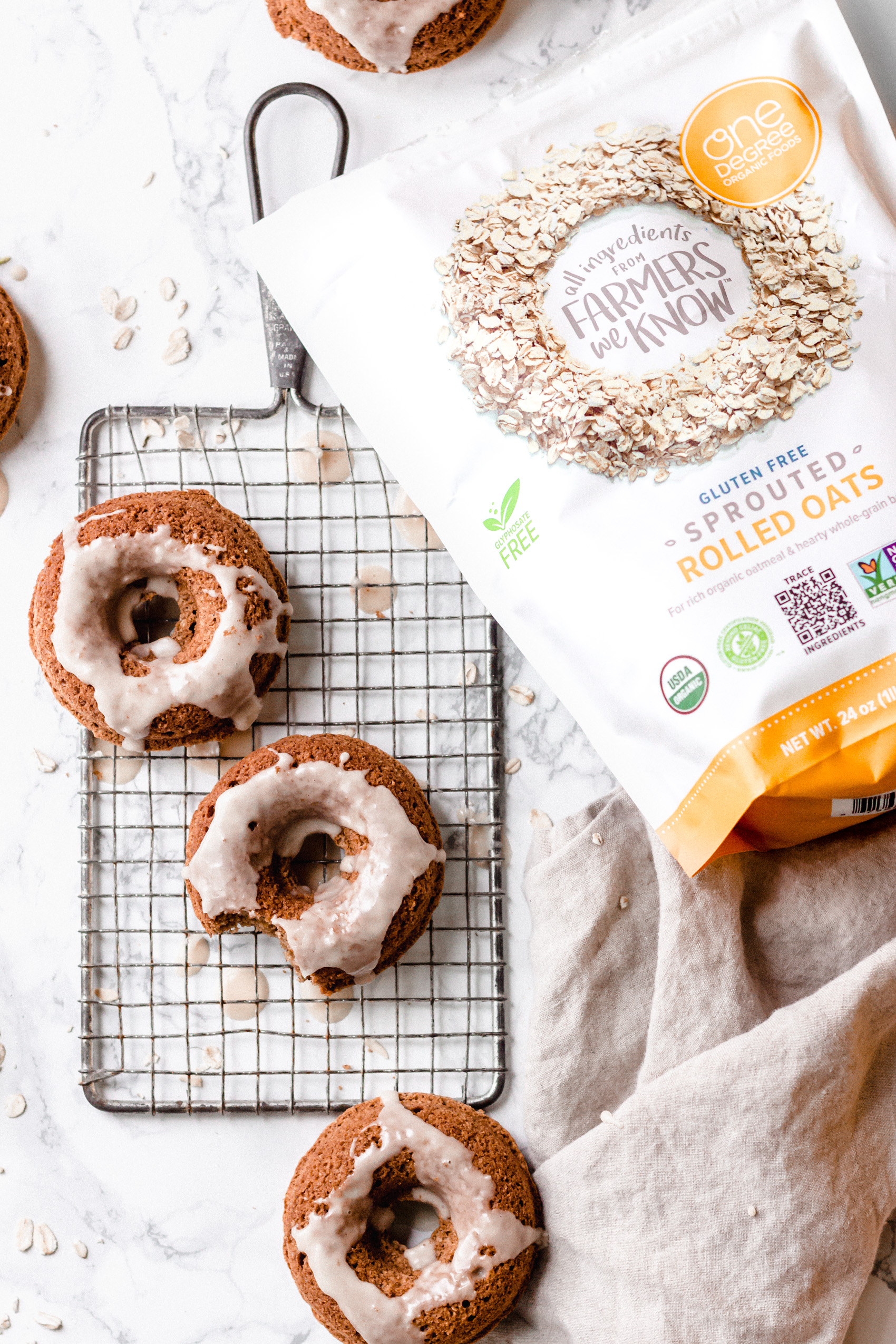 an overhead shot of three glazed baked donuts on a cooling rack next to a bag of sprouted oats and a linen towel