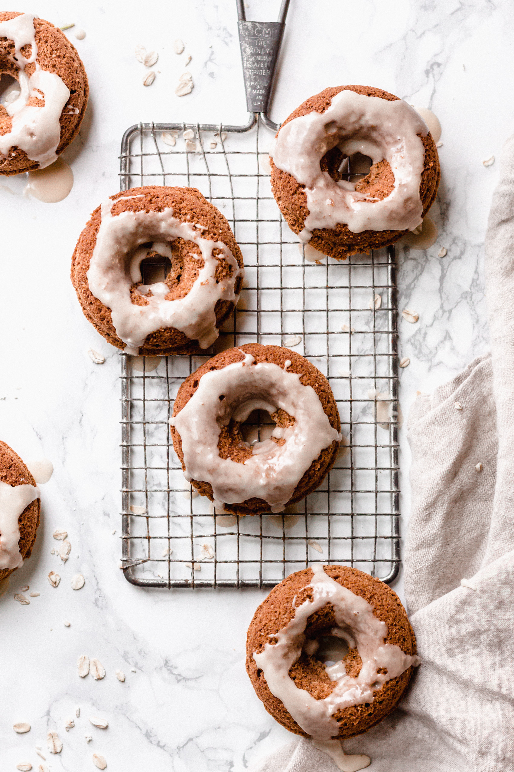 six baked cinnamon donuts, each topped with a maple glaze served on a metal wire serving tray resting on a white counter top next to a linen towel