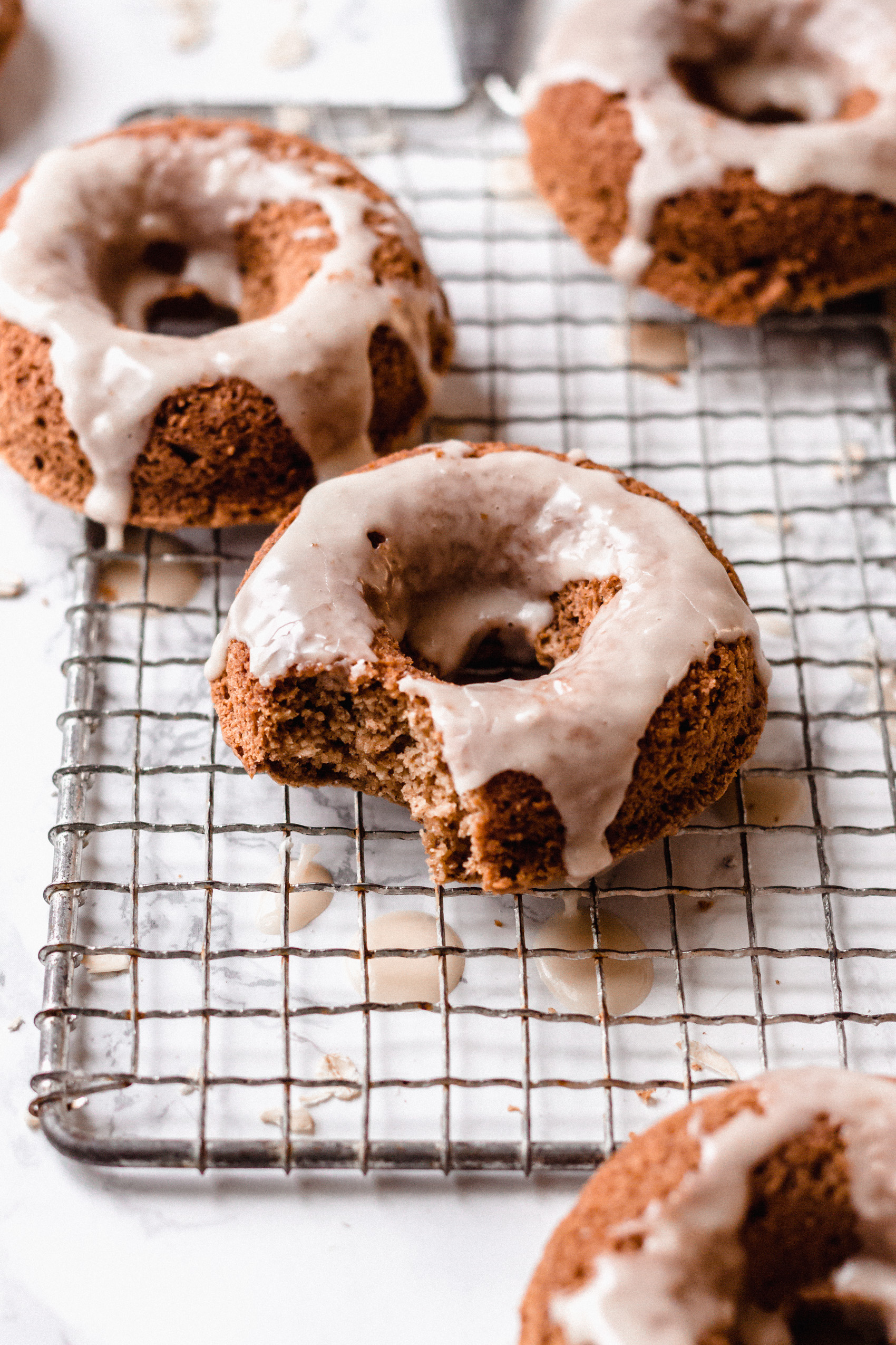 a closeup of a glazed cinnamon baked donut with a bite taken out of it resting next to three other donuts on a wire cooling rack