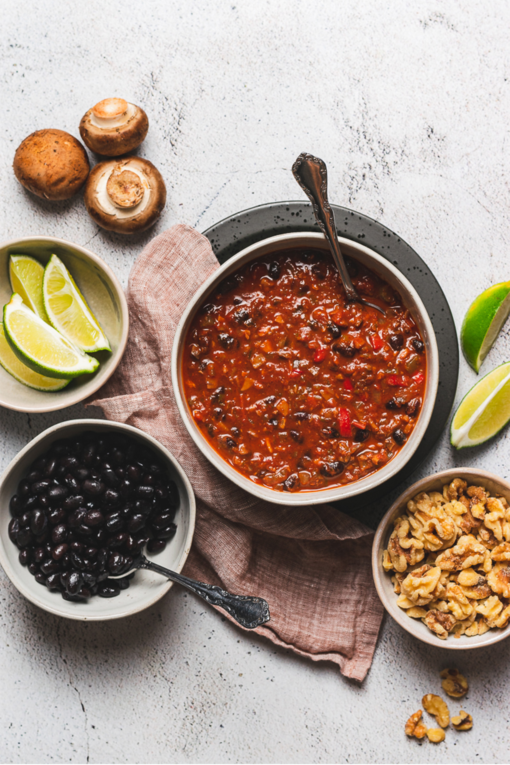 a bowl of vegan chili next to a bowl of limes, black beans and walnuts