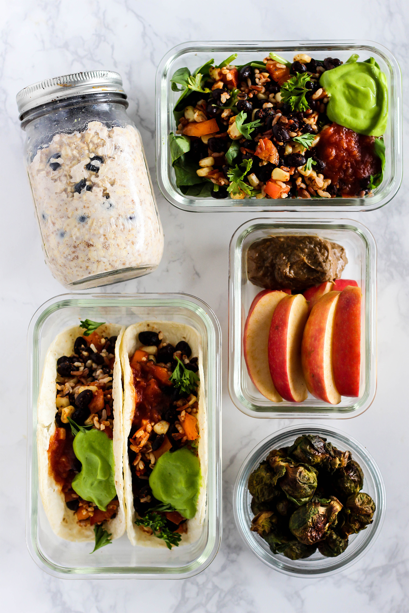 a collection of meal prep containers, one with apple slices and nut butter, one with a salad, one with overnight oats, one with tacos and one with brussels sprouts